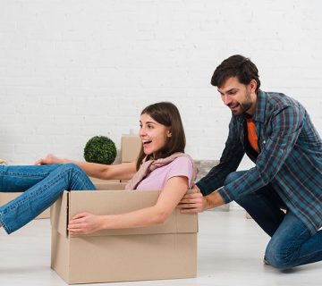 A man pushes a smiling woman sitting inside a cardboard box in a room with a white brick wall and unpacked items.