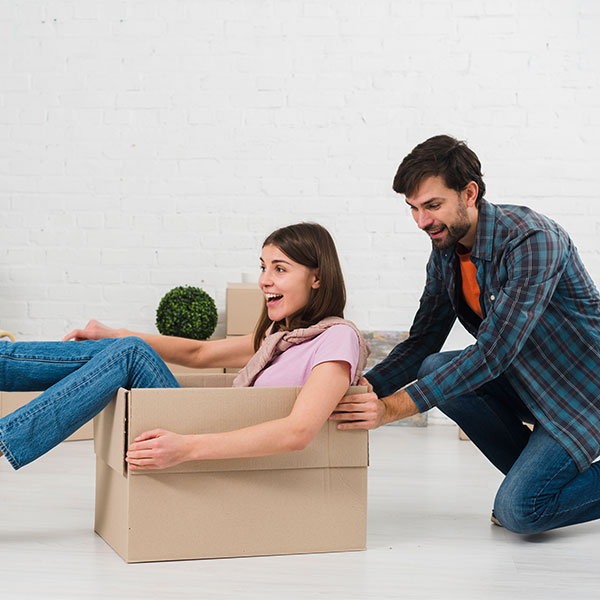 A man pushes a smiling woman sitting inside a cardboard box in a room with a white brick wall and unpacked items.