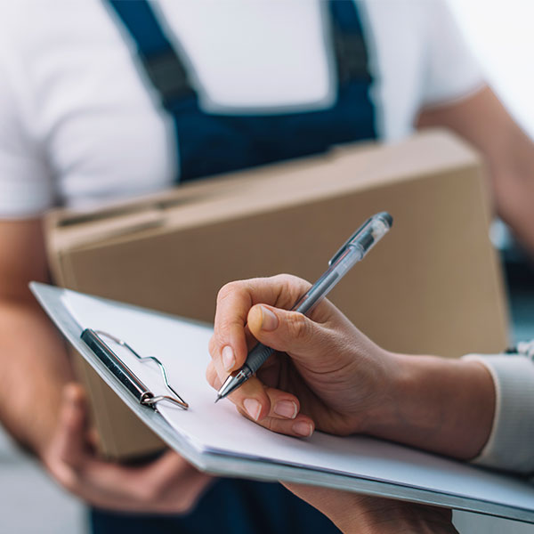 A person signs a clipboard while another person in a uniform holds a cardboard box, suggesting a package delivery or receipt process.