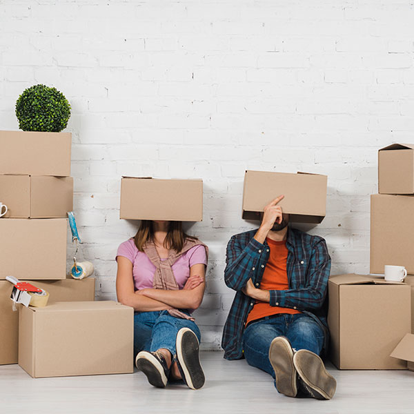 Two people sitting on the floor among moving boxes with cardboard boxes covering their heads.