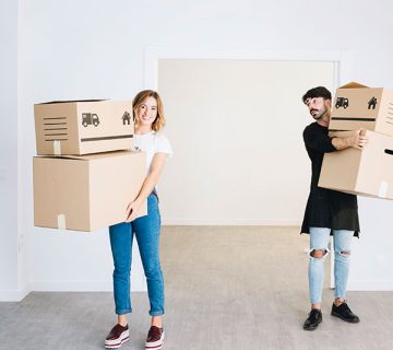 Two people standing in an empty room, each holding cardboard moving boxes and looking in different directions.