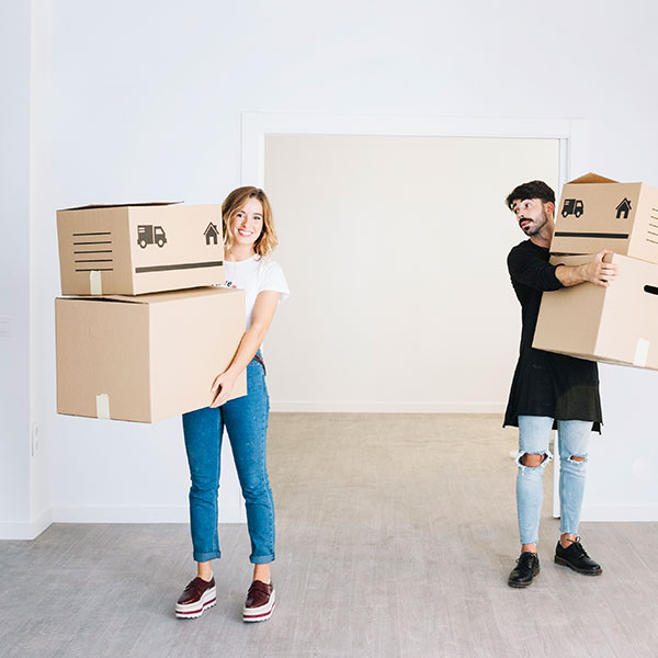 Two people standing in an empty room, each holding cardboard moving boxes and looking in different directions.