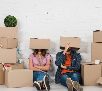 Two people sit on the floor among moving boxes with cardboard boxes on their heads against a white brick wall.
